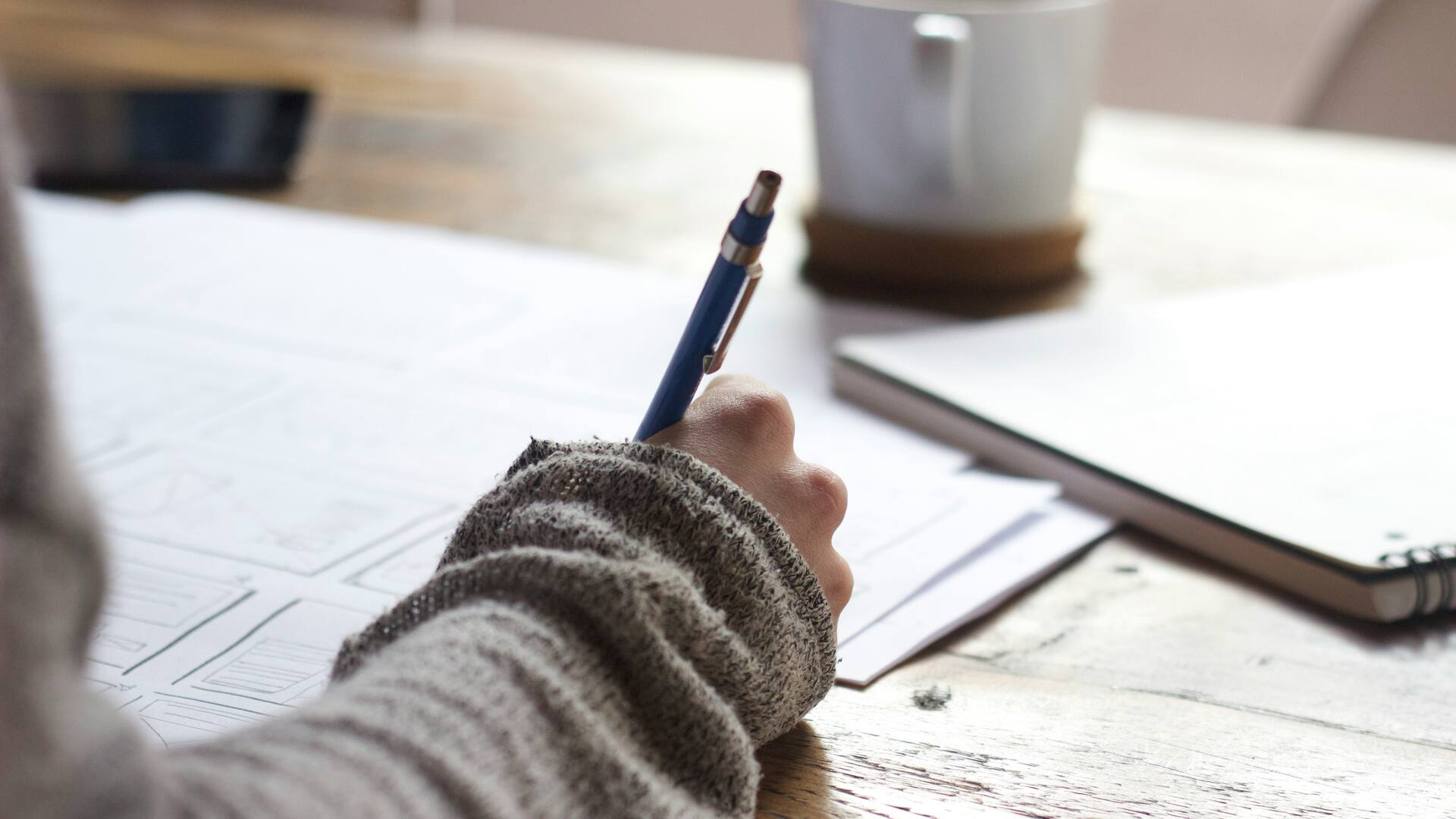 Une main annote un plan au crayon sur une table en bois, à côté d’un carnet et d’une tasse.