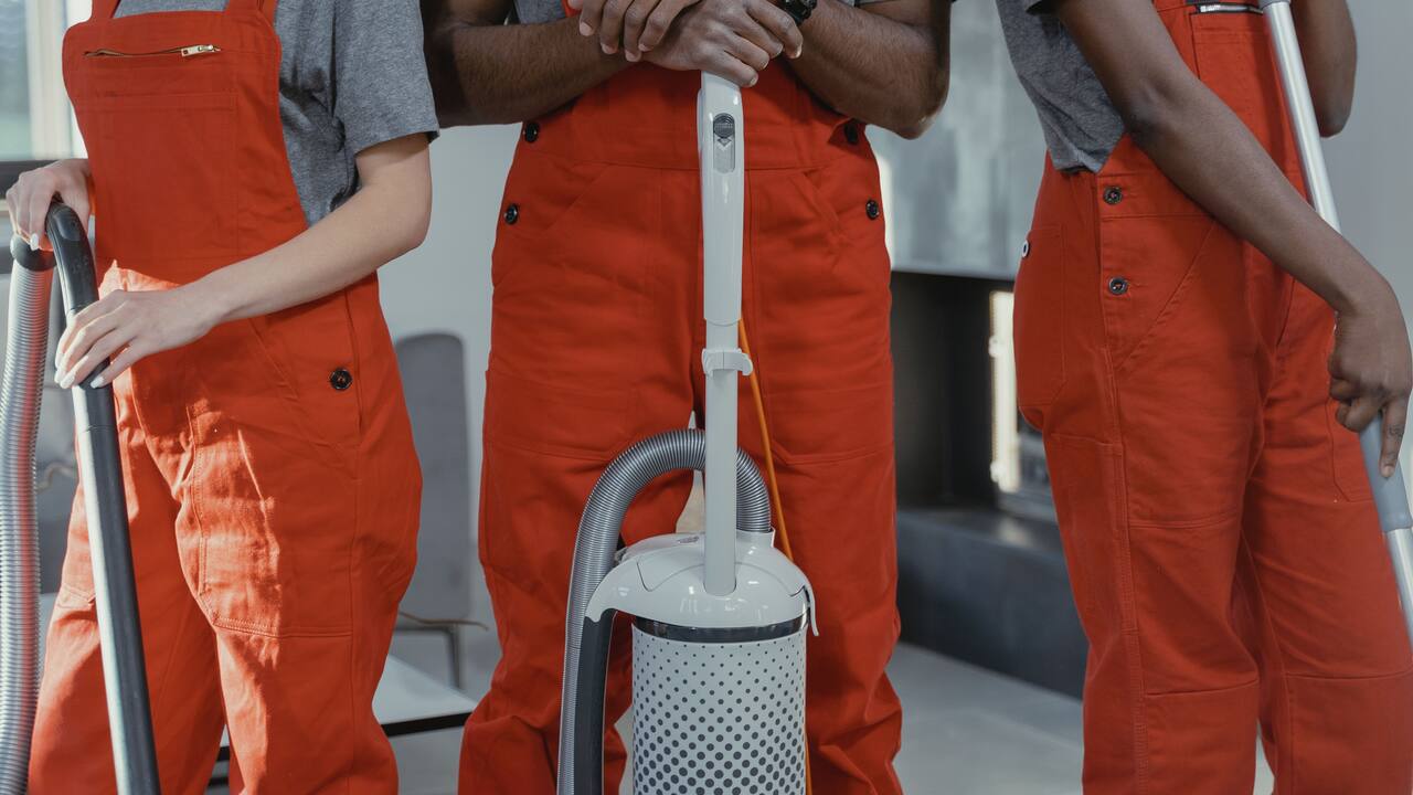 Trois personnes en salopette de travail orange, cadrées au buste, autour d’un aspirateur professionnel et de deux manches de raclette.