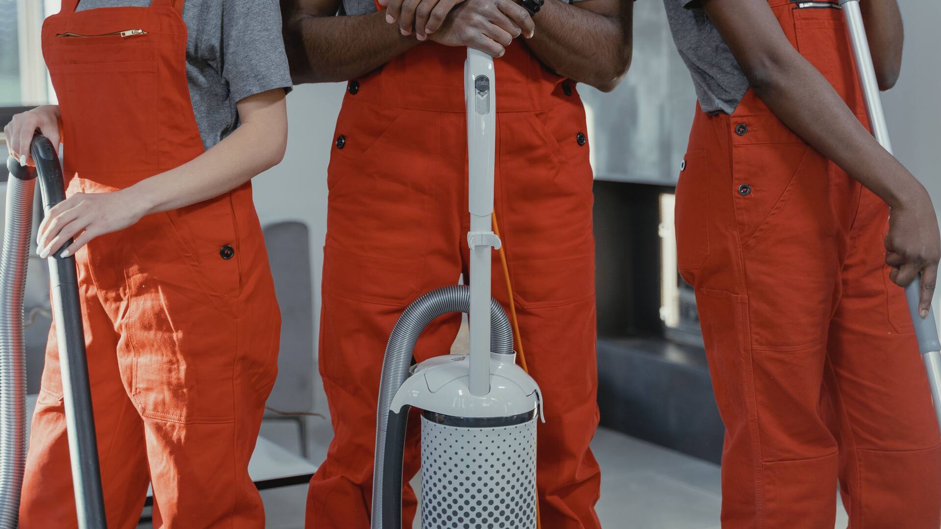 Trois personnes en salopette de travail orange, cadrées au buste, autour d’un aspirateur professionnel et de deux manches de raclette.