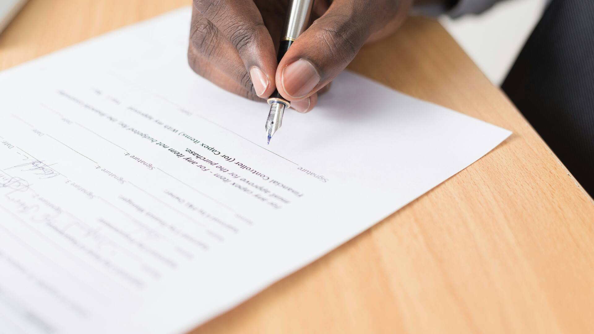 Une main signe un document au stylo plume, sur un plan de travail en bois.