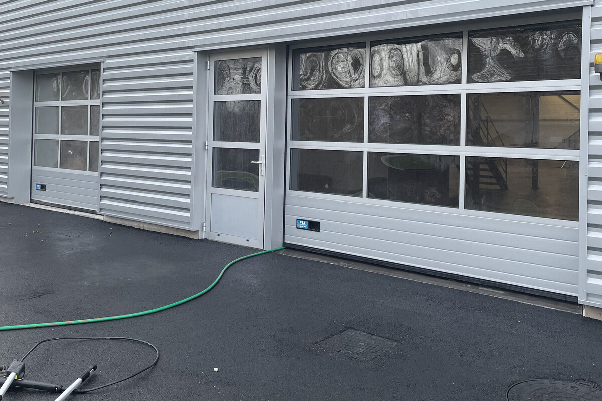 Bardage métallique et portes sectionnelles d’un bâtiment d’activité, tuyau et perche de lavage posés sur l’enrobé mouillé.