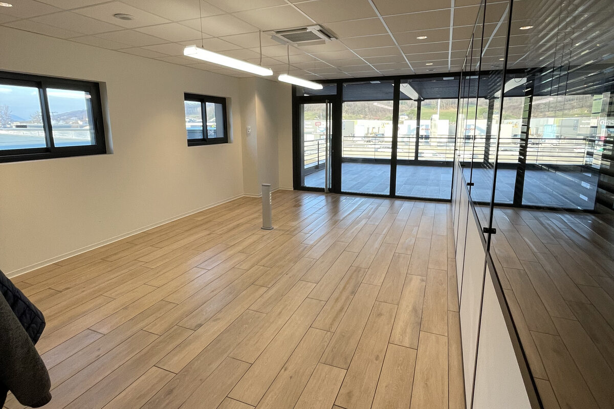 Plateau de bureaux vide après passage, sol en grès cérame imitation bois, cloison vitrée et baie donnant sur une zone d’activité.