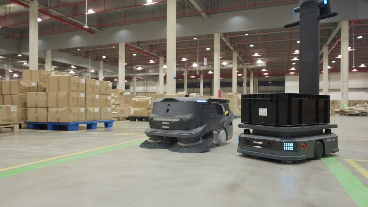 Balayeuse autonome PUDU MT1 et robot de transport T300 dans l’allée centrale d’un entrepôt, devant des palettes de cartons.