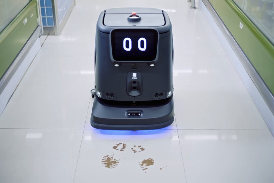 Robot autolaveur PUDU CC1 Pro effaçant des traces de chaussures sur le carrelage blanc d’une circulation vitrée.