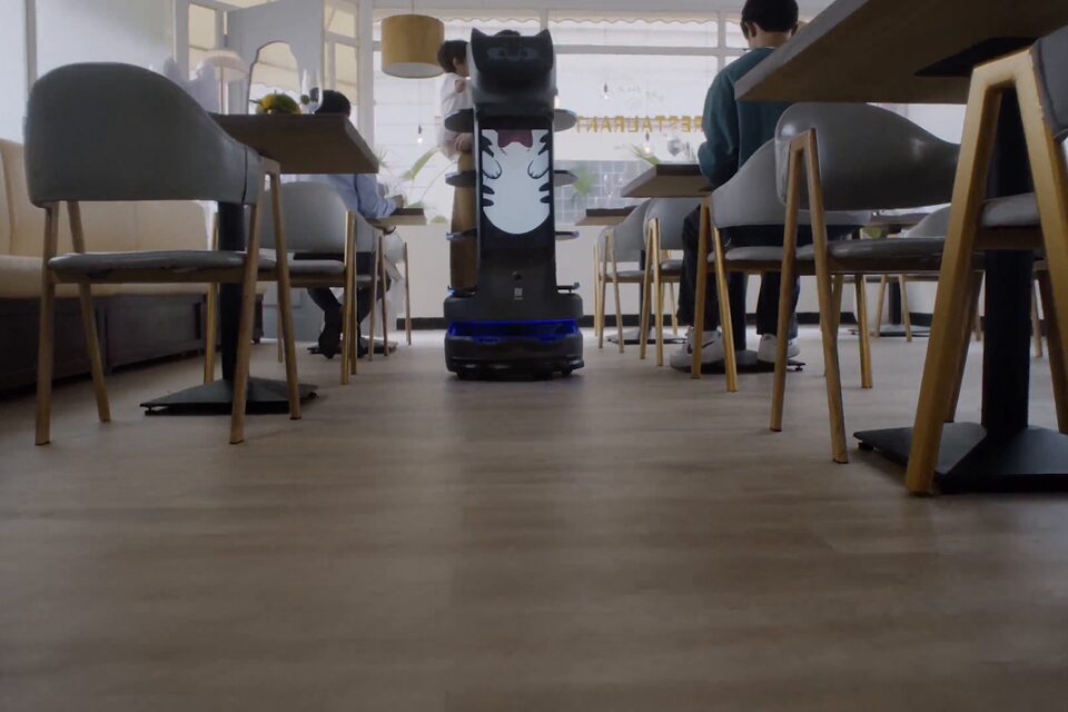 Robot de service PUDU BellaBot Pro à l’arrêt entre les tables d’une salle de restauration collective.