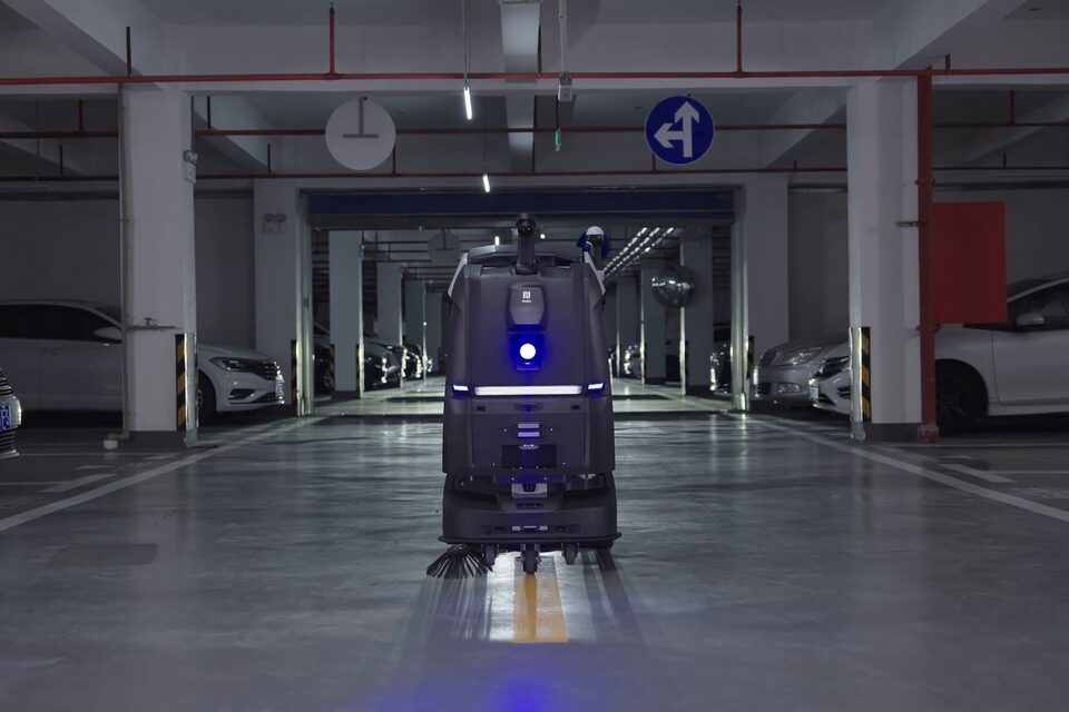 Robot autolaveur PUDU BG1 Pro balayant l’allée d’un parking souterrain, entre deux rangées de véhicules.