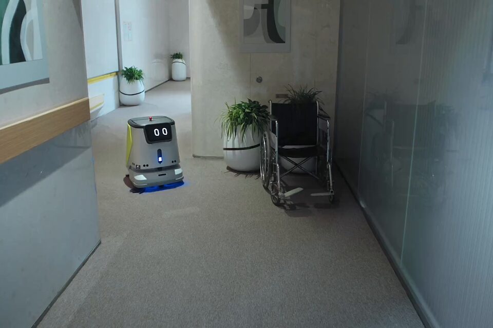 Autolaveuse autonome de la série PUDU CC1 remontant la circulation d’un établissement de soins, trace de lavage visible derrière elle.