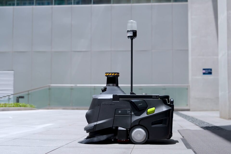 Robot balayeur PUDU MT1 Max sur le parvis extérieur d’un bâtiment, le long d’une façade vitrée.