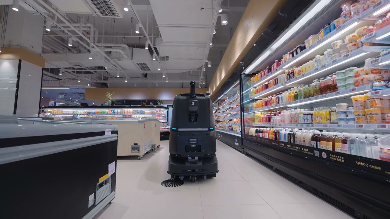 Autolaveuse PUDU BG1 Pro remontant l’allée carrelée d’un supermarché, les meubles réfrigérés éclairés sur toute la longueur.