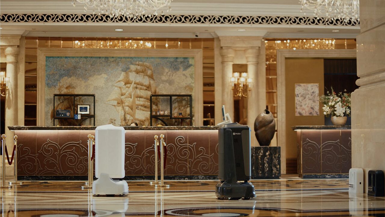 Deux robots de service PUDU à l’arrêt dans le lobby d’un hôtel, devant le comptoir de réception et son sol de marbre.
