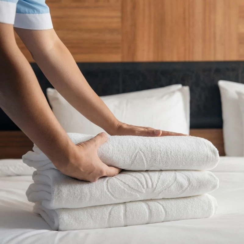 Agent de conciergerie pliant des serviettes blanches impeccables sur un lit pour la préparation d'un logement Airbnb à Annecy.