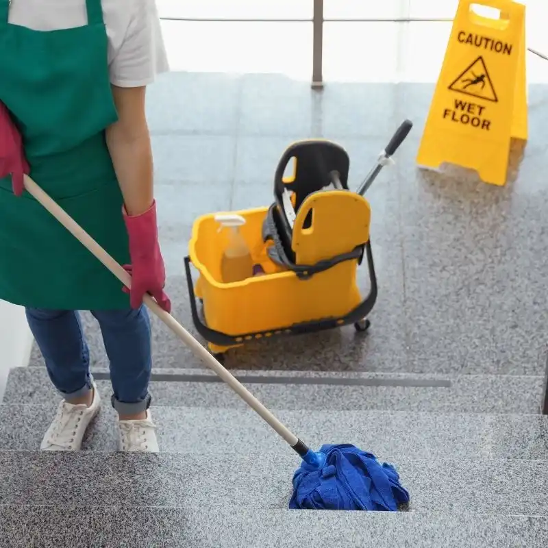 Agent de propreté passant la serpillière dans les escaliers d'une copropriété en Haute-Savoie, avec panneau d'avertissement de sol glissant.