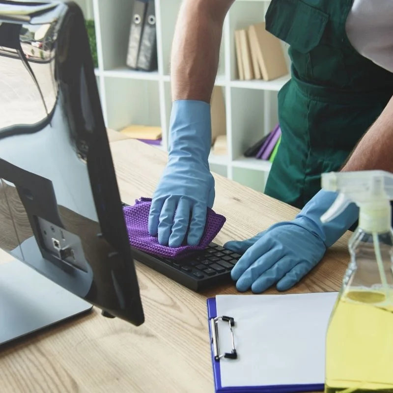 Agent d'entretien avec gants bleus nettoyant minutieusement un bureau et un clavier d'ordinateur professionnel à Annecy.